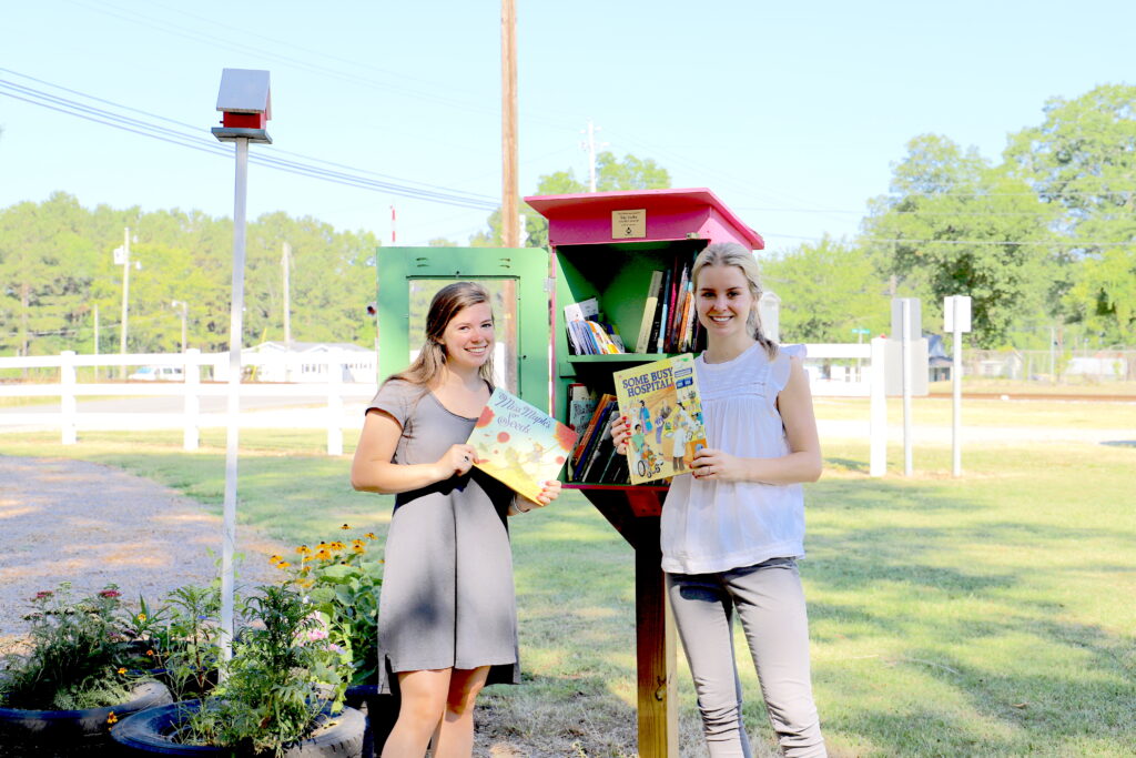 Two young women stand in front of a little lending library for books in a park. They are each holding a book and smiling. 